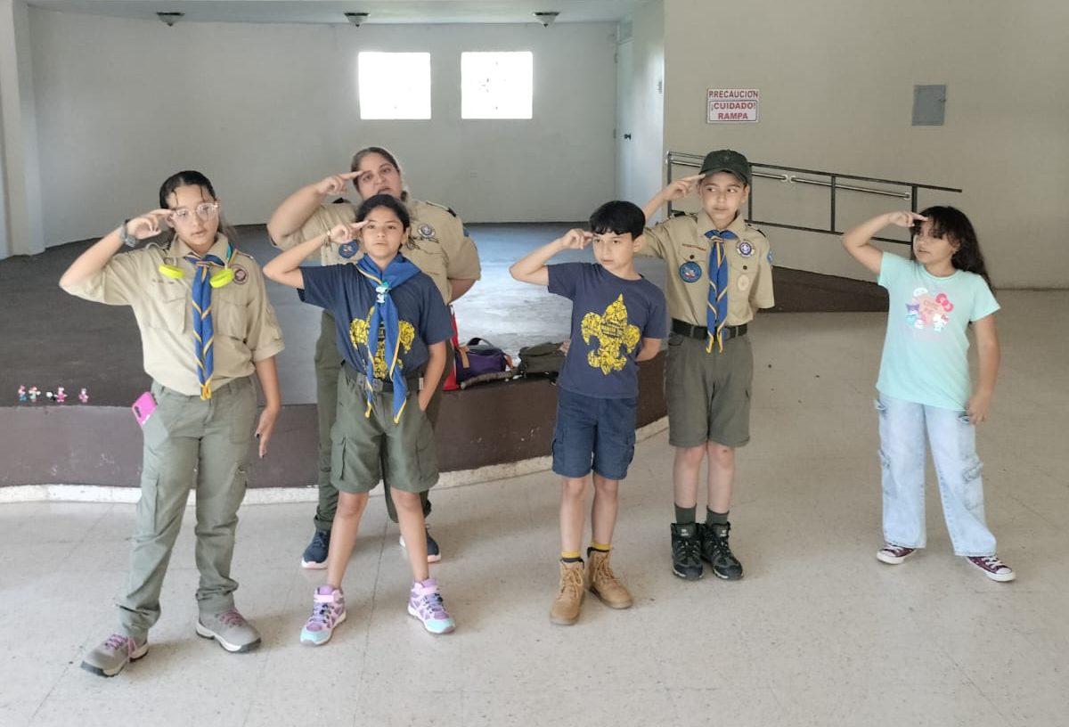 5 cobitos en línea uno al lado del otro haciendo el saludo a la bandera, con su mano derecha colocando dos dedos (índice y medio) sobre la ceja derecha. Su líder Jenny, está justo detrás de ellos habiendo el saludo.